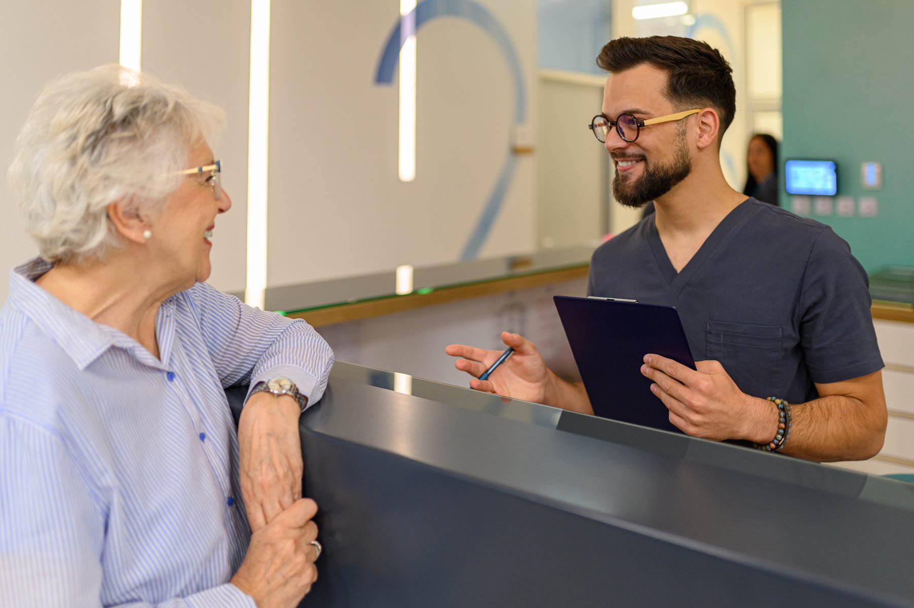 Front desk team member with a clipboard, smiling and talking with an older patient at the check-in counter.
