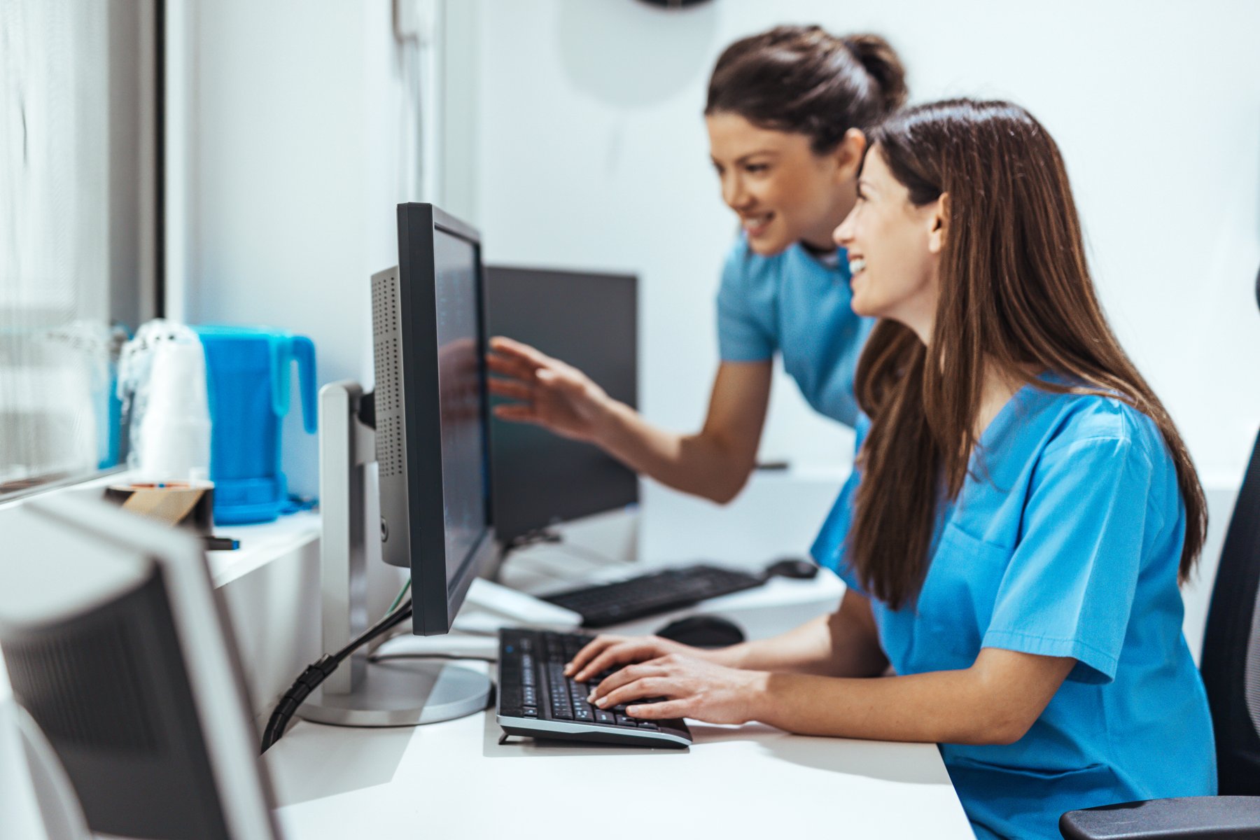 Two team members in scrubs smiling and working together at a computer.